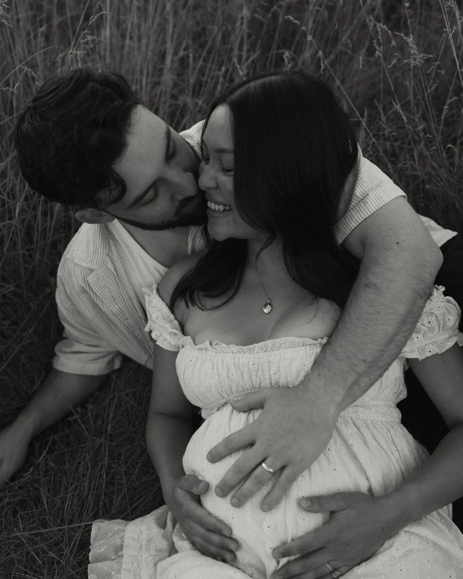 A black and white portrait of Angela and Josh cuddling in long grass