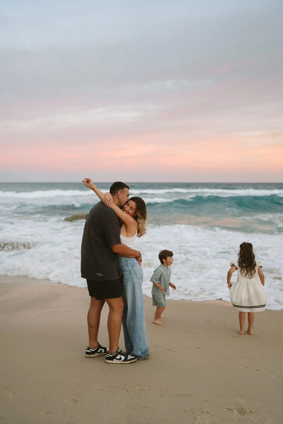 Parents kissing on the sand at sunset while their kids play nearby