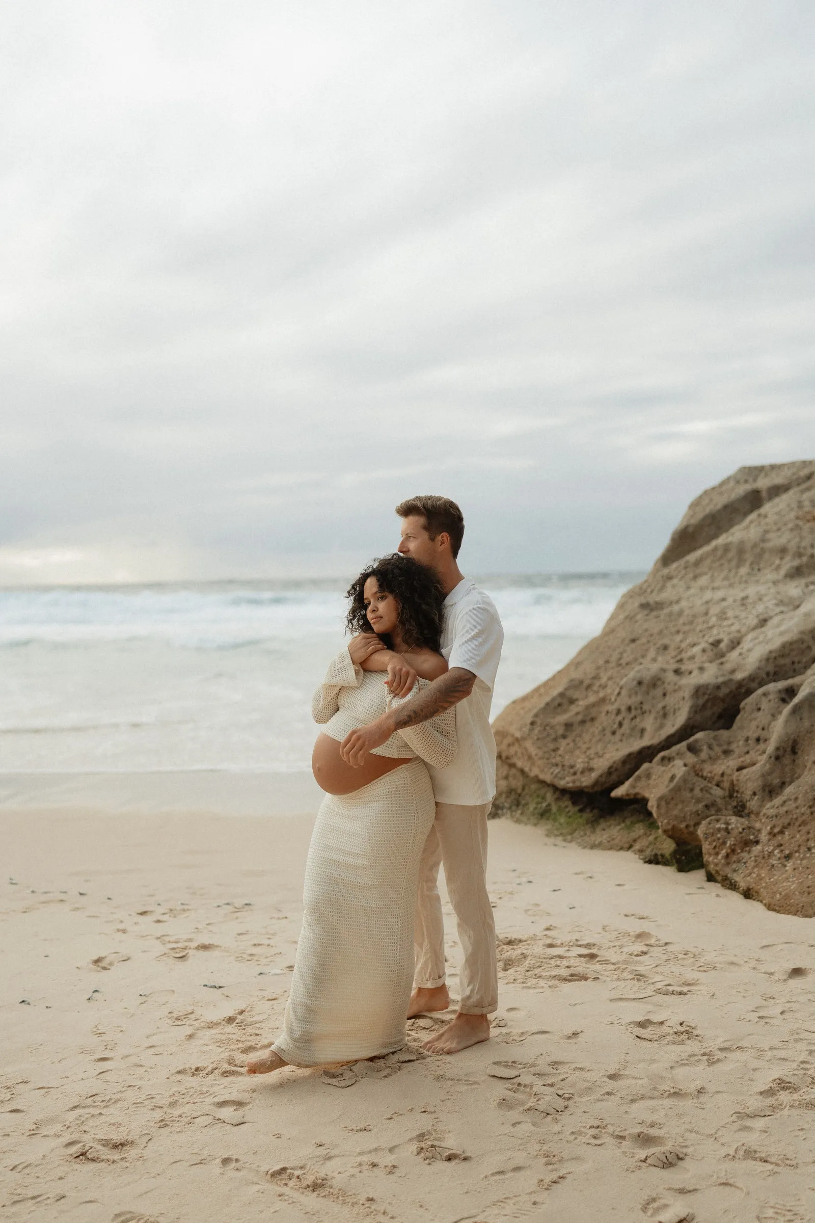 An expecting couple embracing on the beach beside sandstone rocks