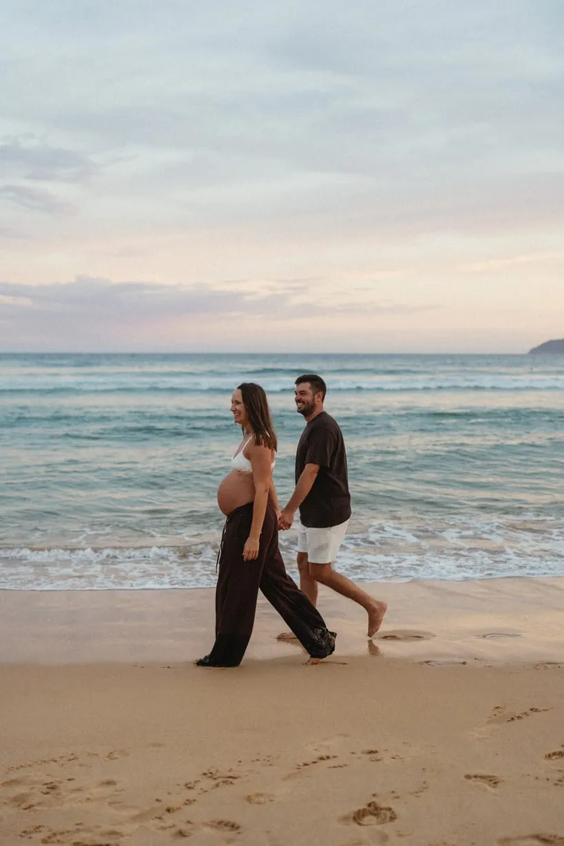 An expecting couple walking along the shoreline at dusk
