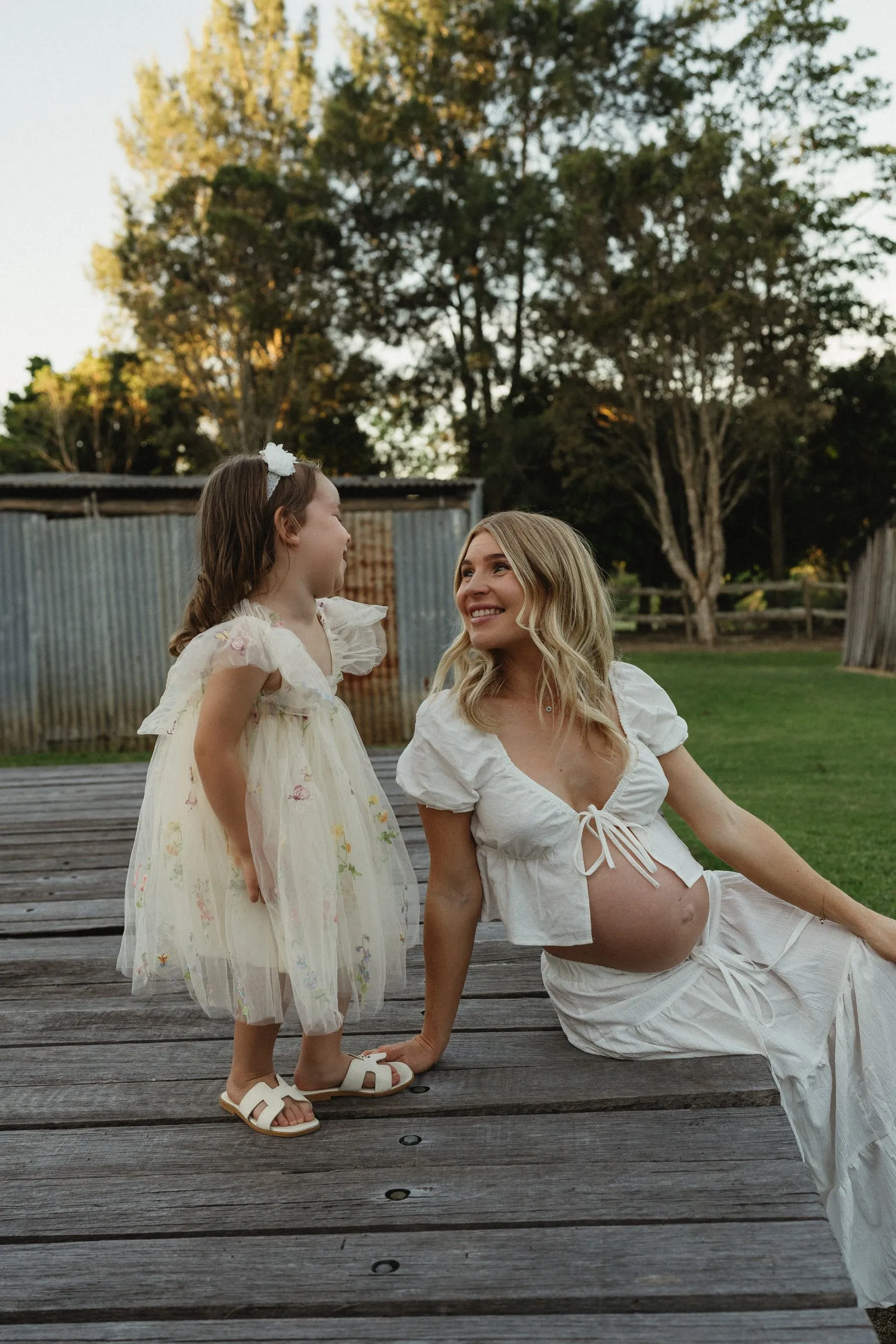 An expecting mum and her daughter smiling at each other on a garden bench