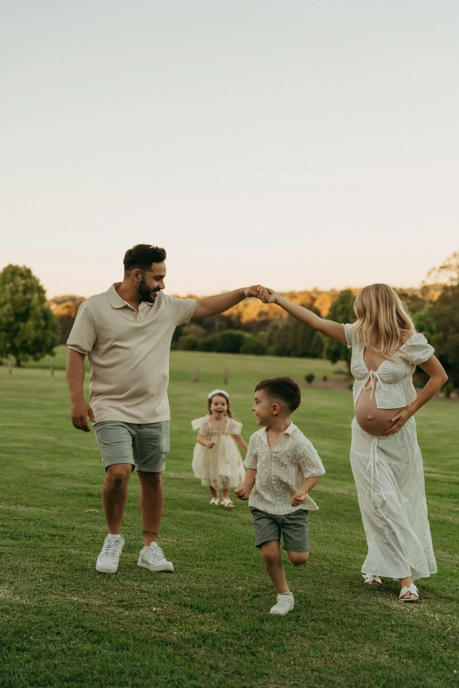 An expecting mum twirling with dad while their children play in the grass