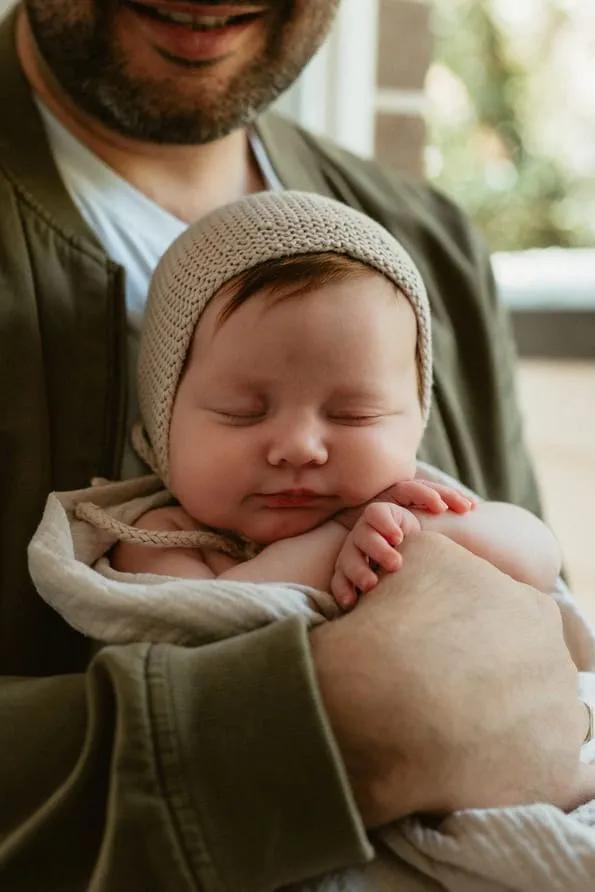 A sleeping newborn in a knitted bonnet held in dad’s arms
