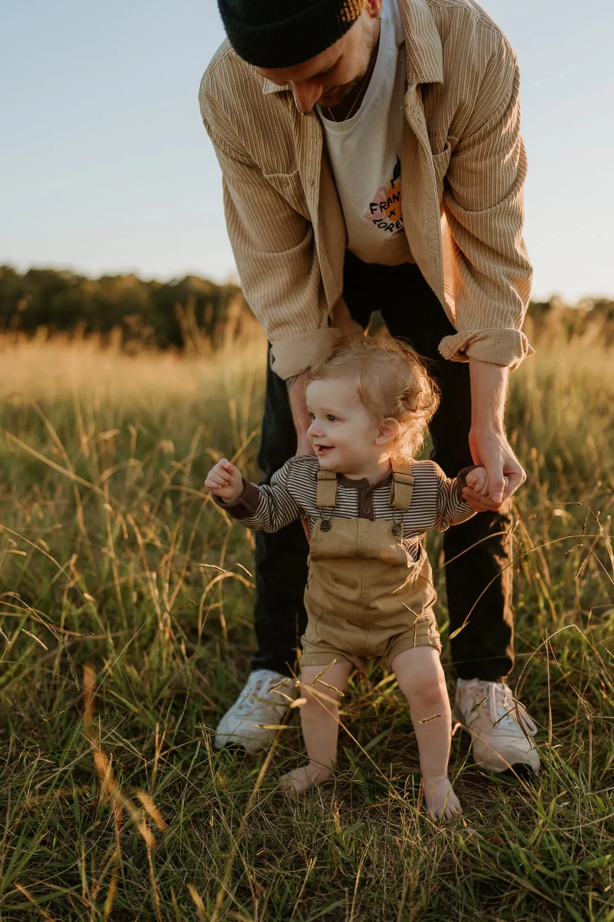 A dad holding his toddler’s hands as he walks through tall grass
